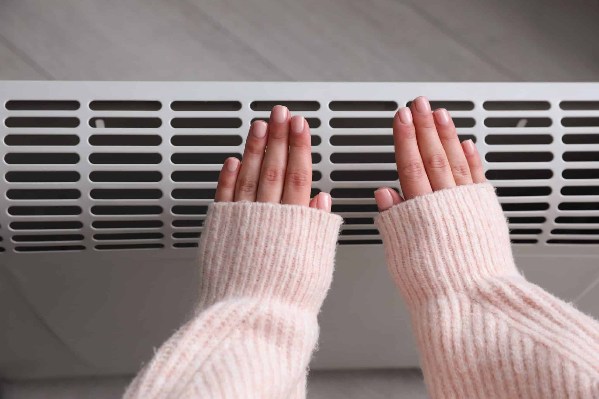 A person wearing a pink sweater warming their hands over an electric heat pump with a white floor background.