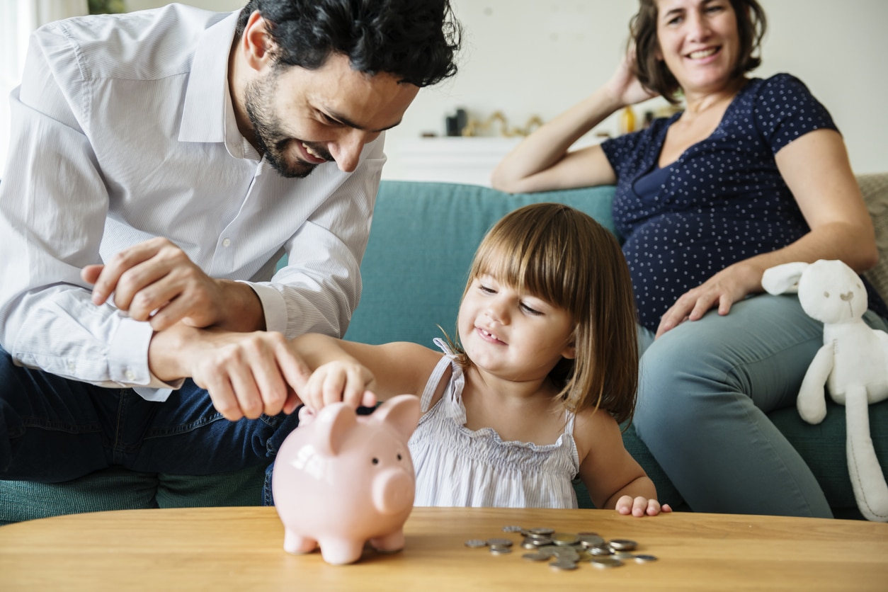 A mother, father, & daughter sitting in a living room smiling while the daughter puts money into her piggy bank.