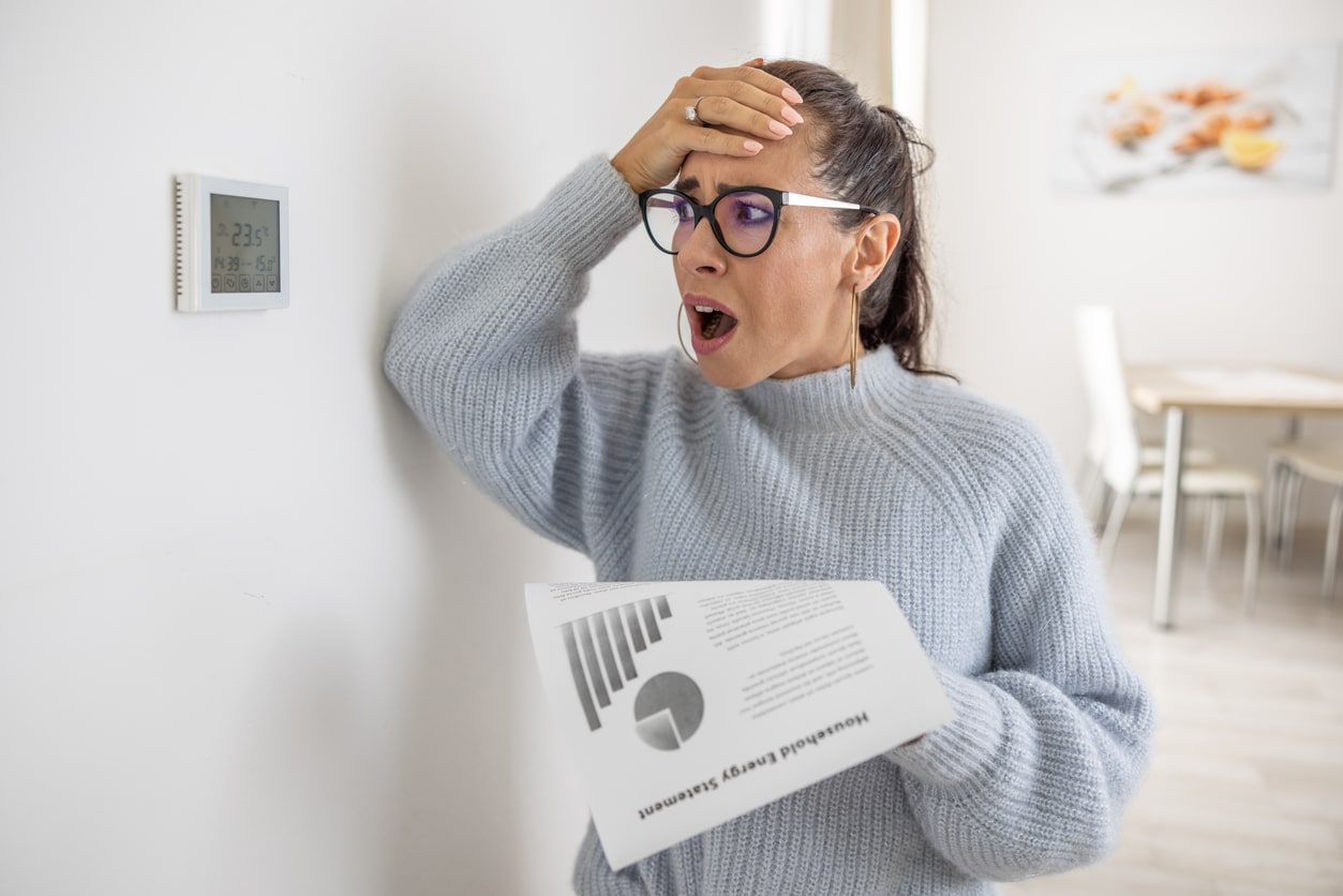 woman wearing a gray sweater looking at her thermostat wishing she could save money on heating.