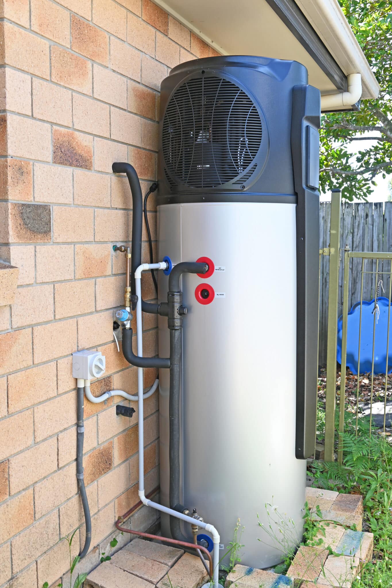 A silver and blue heat pump water heater outside of a tan brick home.