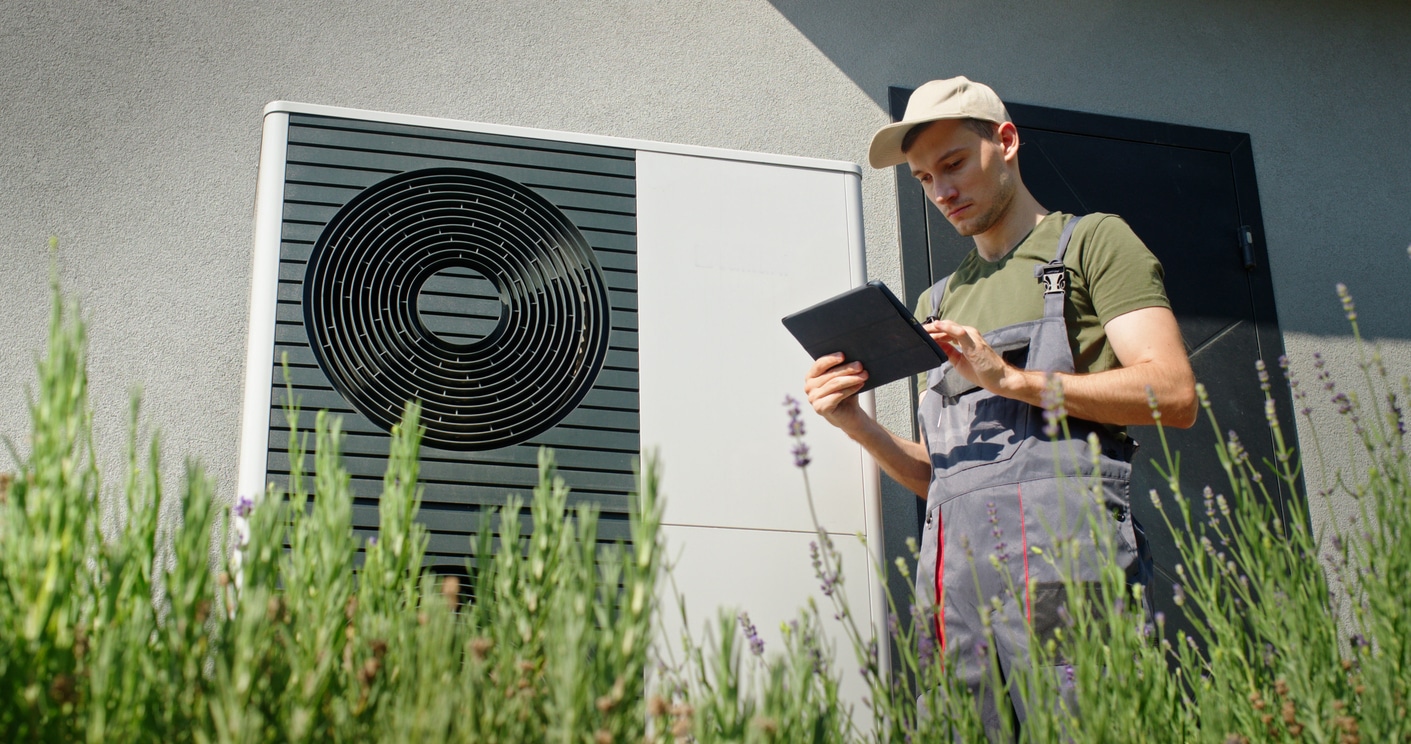 A young technician in overalls and a cap inspects an outdoor heat pump system using a tablet.