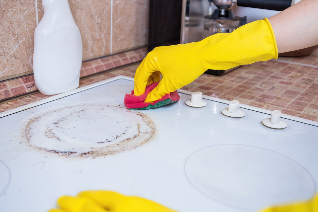 A person wearing bright yellow rubber gloves cleaning a brown stain on a white electric stove top.